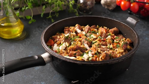 A hand placing a hot frying pan with a Mediterranean-style meat and vegetable dish onto a metal trivet. Dark kitchen countertop background with fresh herbs, olive oil, and cherry tomatoes in 4K.