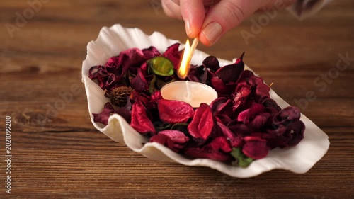 Close-up of a hand lighting a small tea light candle placed inside a white decorative shell filled with vibrant pink and red dried flower petals. The scene is set on a rustic wooden table, evoking a s
