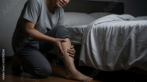 A person suffering from a painful leg cramp or muscle spasm in their calf while kneeling on the bedroom floor next to a bed.