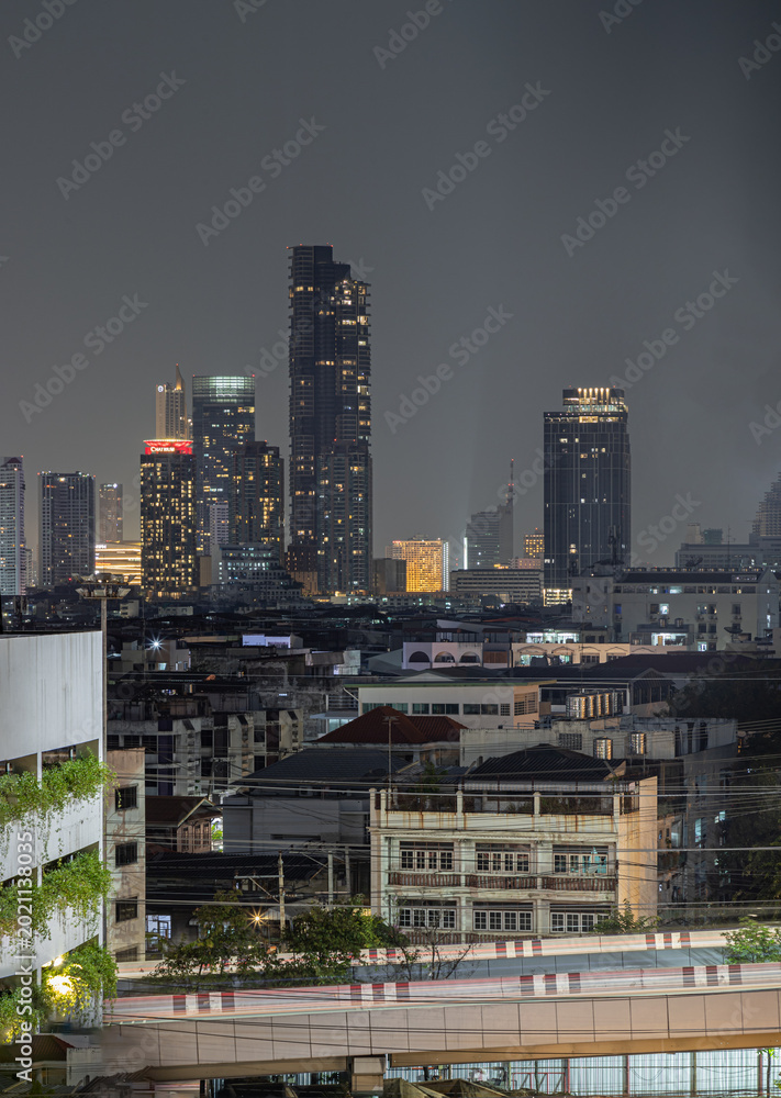Obraz premium A cluster of gleaming skyscrapers pierces the heavy twilight sky, casting a brilliant array of golden and white lights over the expansive cityscape below. In the foreground, a tapestry of low-rise nei