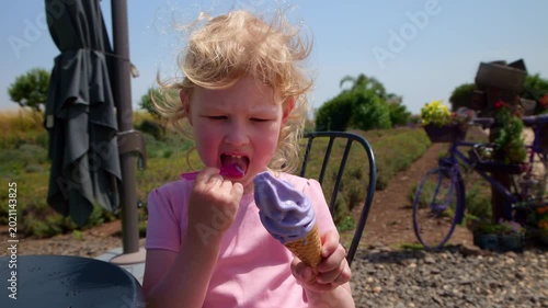 A little blonde girl with curly hair, wearing a pink dress, sits at an outdoor cafe table and eats purple lavender ice cream with a small spoon. In the background is a lavender farm