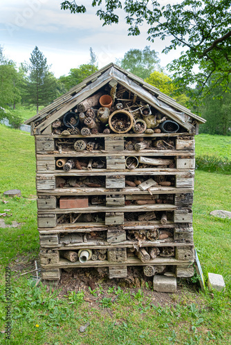 A handmade insect hotel built from weathered wood, pinecones, and hollow stems stands in a green park beneath leafy summer trees.