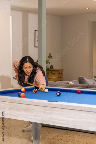 Asian woman aiming pool shot on blue-felt table with cue-stick at home in pink sweater