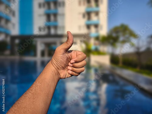 Close-up shot of a person's hand giving a thumbs-up gesture in front of a blurred background with a swimming pool and apartment buildings on a sunny day.
