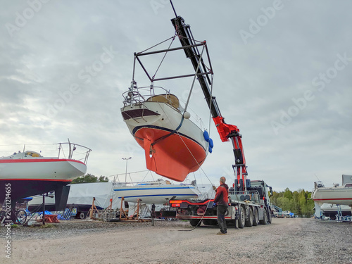 Obraz na płótnie A white sailboat with a red hull is hoisted into the air by a hydraulic crane on
