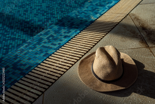 Fotografía Straw hat on poolside stone deck with blue swimming pool water and sunlight shad