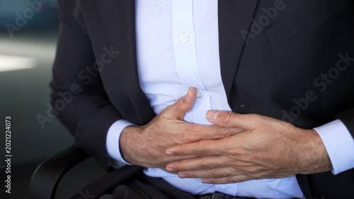 Close up. Businessman in formal suit feels stomach pain sitting at workplace in business office. Male hands holding massaging stomach. Sick man worker suffers from gastritis, poisoning or bloating