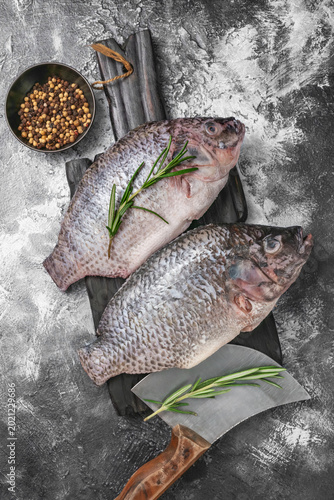 Two fresh raw tilapia fishes on a dark wooden board with rosemary and peppermorns.