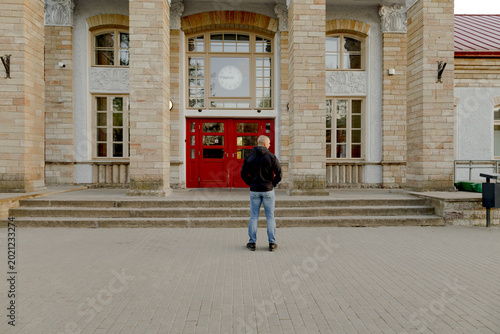 Man standing in front of historic stone building with red entrance doors and large clock during sunny day in urban environment.