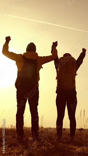 A man and woman stand together, raising their arms in celebration at sunset. The couple enjoys the view, expressing joy against the backdrop of the sun.