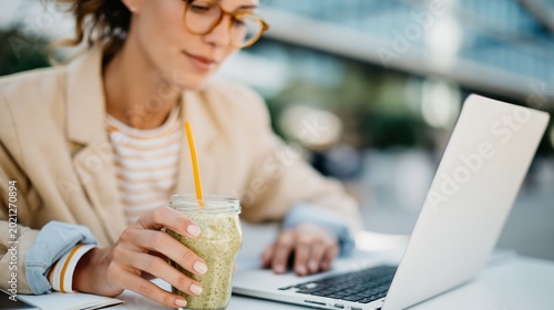 A professional, stylish woman sipping a green smoothie while working on her laptop outdoors, blending productivity with healthy living in a modern workspace.