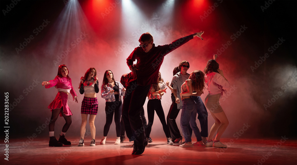 Fototapeta premium Male lead dancer performing in front of energetic group under red stage lights. Concept of dance crew promo, live show poster, music festival visuals and nightlife campaign.