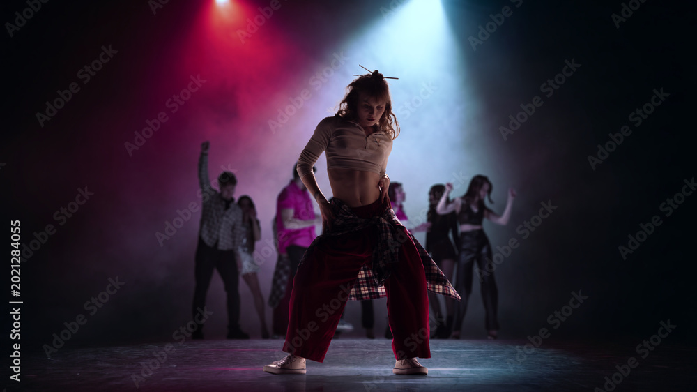 Naklejka premium Female street dancer posing under blue spotlight with dance crew in background. Concept of dance school ad, performer casting, youth training promo and urban event visuals.