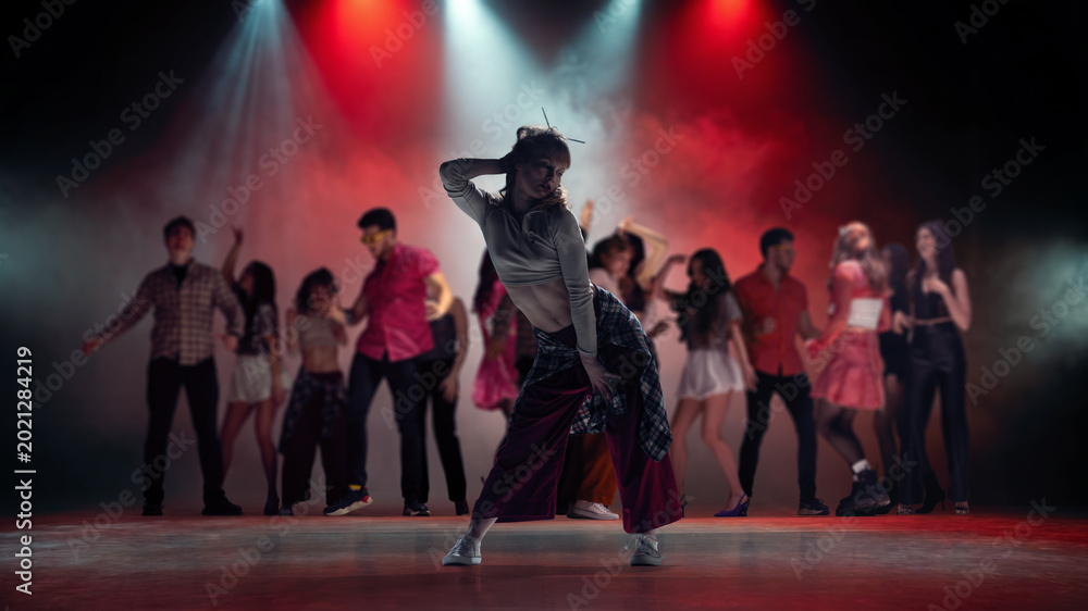 Fototapeta premium Young female dancer posing in smoky concert lights with group dancing behind. Concept of dance class campaign, stage show poster, music video casting and youth culture promo.