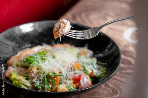 Fényképezés A fork lifts a piece of shrimp covered in sauce from a gourmet Caesar salad topped with shredded parmesan cheese in a black bowl