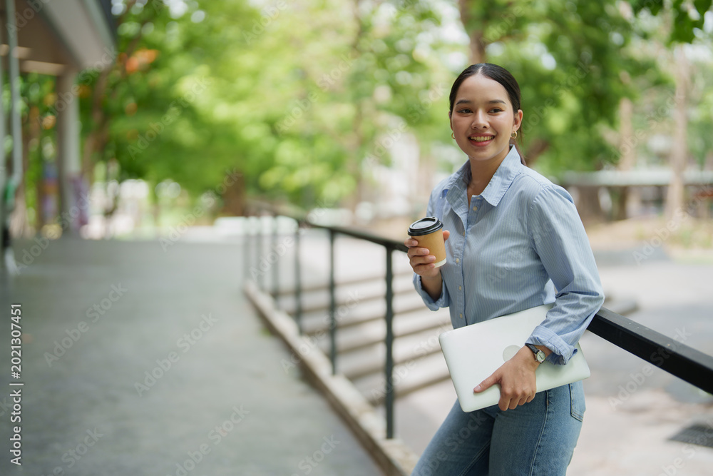 Fototapeta premium Asian university student or young freelancer smiling with a positive expression, holding a laptop and a takeaway coffee cup outdoors on a university campus or modern workspace area