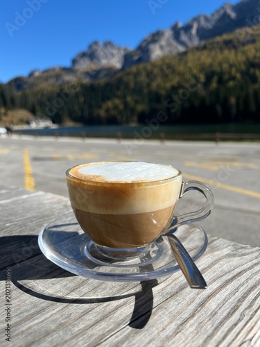 Cup of cappuccino on a wooden table with Lake Misurina and the Dolomite mountains in the background. Calm autumn morning with sunlight and mountain reflections.
