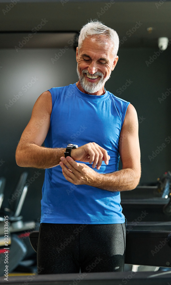 Obraz premium Portrait of a senior man exercising in a gym, mature male running using treadmill machine and checking smart watch for performance, healthy lifestyle and cardio exercise at fitness club concepts, vita