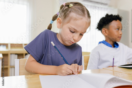 Photos Two children in a classroom, one focused on writing in a notebook, the other looking to the side, sitting at a wooden desk with school supplies