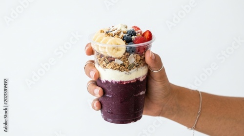 Hand holding a layered acai bowl in a clear plastic cup with fruit and granola