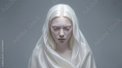 Albino woman with long white hair wearing silk veil. Ethereal portrait of young female looking down with sad expression against gray background.