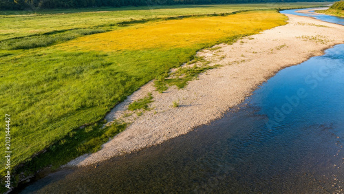 Aerial view of a riverbank with green and yellow grasslands adjacent to calm blue water
