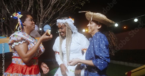 Three happy Brazilian women in traditional Festa Junina costumes celebrate the June festival at night, laughing and eating typical snacks while wearing colorful caipira and bridal outfits