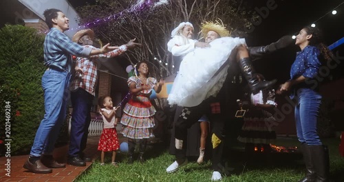People celebrating a traditional Brazilian Festa Junina at night, featuring a mock wedding ceremony where a man lifts a bride in a white dress while guests in caipira costumes cheer and dance