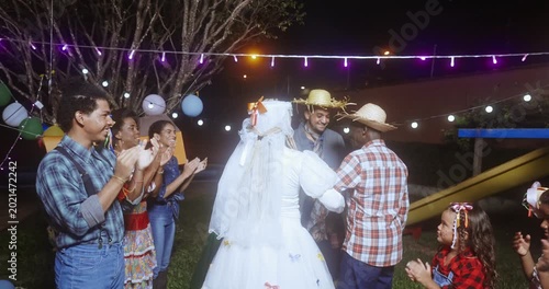 A joyful group of people celebrates a traditional Brazilian Festa Junina mock wedding ceremony at night, featuring a bride and groom in typical caipira costumes under colorful festive lights