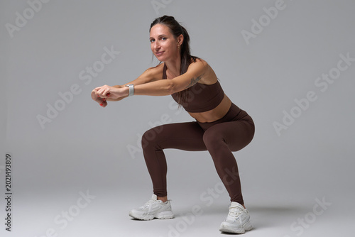 Fit woman doing squatting exercise looking at camera on gray background
