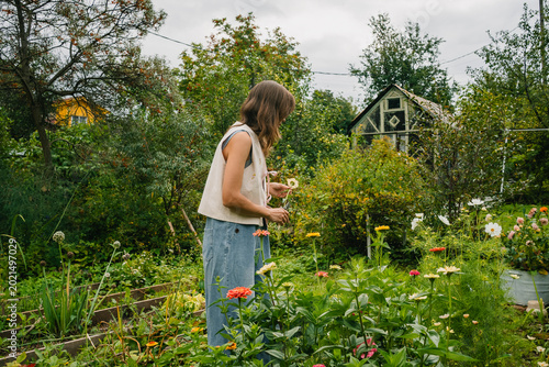 Konstfotografi Woman wearing denim overalls and a cream vest is joyfully cutting vibrant flower