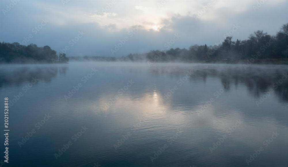 Fototapeta premium Misty morning drifts over calm lake surface. Soft light glows through hazy sky above trees. Reflections ripple gently across still water. Fog curls low near shoreline, veiling distant shore