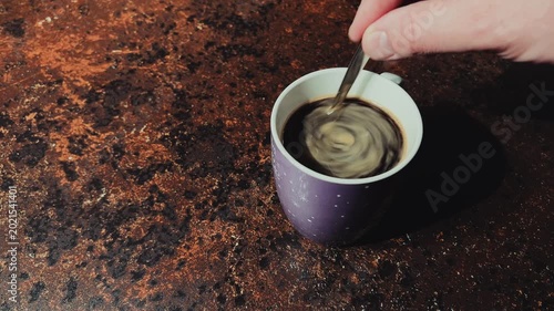 A hand stirs black instant coffee with a metal spoon in a purple ceramic mug. Close-up shot on a dark brown textured surface in natural light.