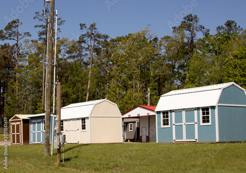 a line of colorful display sheds and outbuildings on a grassy lot with a treeline and utility pole under a clear blue sky