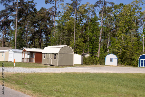 several prefabricated storage sheds and small portable outbuildings arranged on a gravel lot bordered by green grass and tall pine trees. 