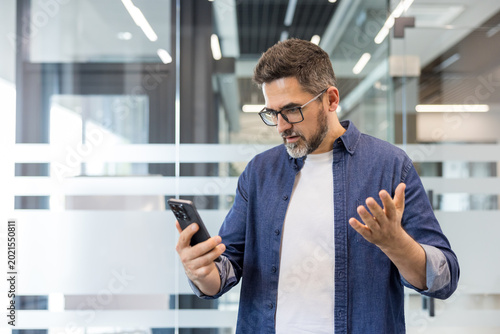 Mature professional holding smartphone, looking frustrated and confused after reading bad news or an upsetting message in a modern office environment, stressed and pensive