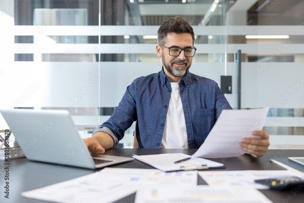 Obraz premium Professional man wearing glasses and a denim shirt sitting at his office desk, happily engaged in work while using a laptop and reviewing financial documents with graphs on the table