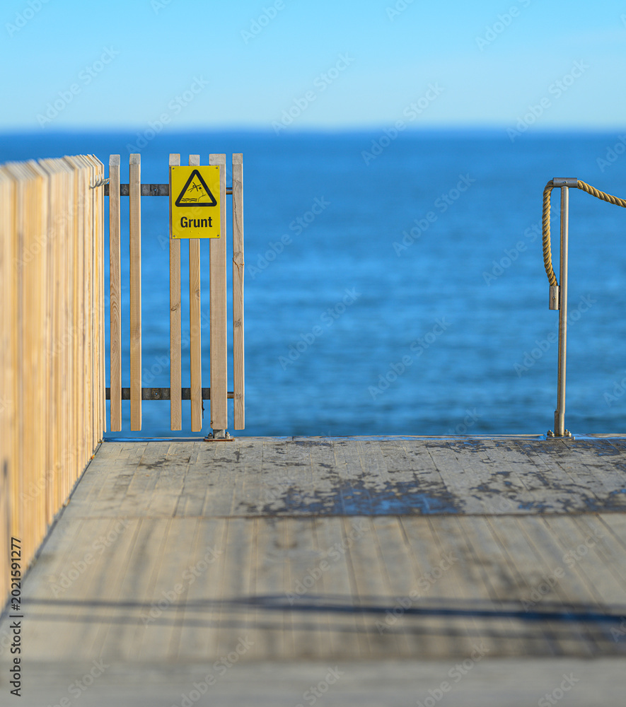 Obraz premium End of pier with safety sign warning of diving into shallow water against calm sea backdrop.