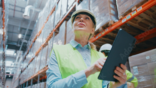 Female warehouse employee walking through warehouse. Holding tablet. Managing logistics or checking inventory data during work process.