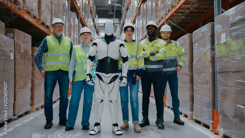 Warehouse staff standing proudly with humanoid robot in storage aisle. Wearing protective gear. Collaboration between humans and AI in logistics environment. Modern distribution center.
