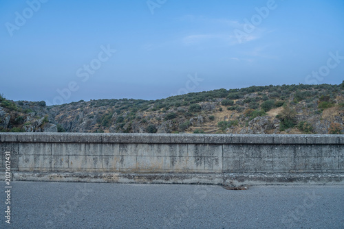Konstfotografi A weathered stone bridge parapet overlooks a paved road, with rocky hills and scattered vegetation under a cloudy blue sky
