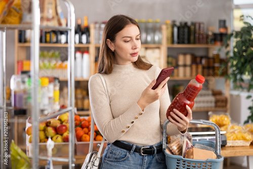 Fotografie Young woman buyer scanning qr code for fresh tomato juice in bottle in grocery s