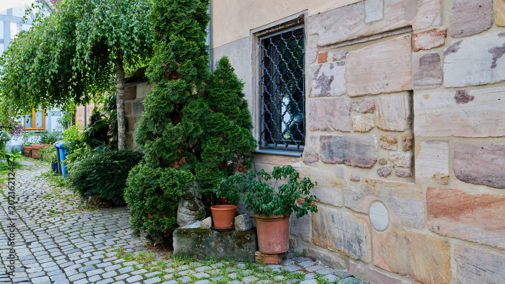 Fototapeta premium Cozy courtyard of an old German house with a rustic sandstone wall, small window with iron bars, green thuja trees, and a cobblestone pathway.