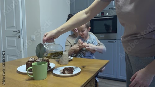 Woman pours tea into cups while mother and child play clapping game at table cozy family moment.