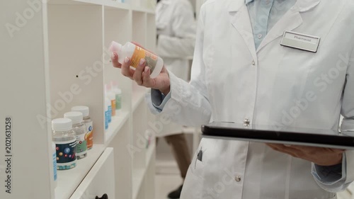 Tilt up detail shot of Caucasian young woman wearing white coat and using tablet while doing stock inventory in pharmacy storage room, copy space