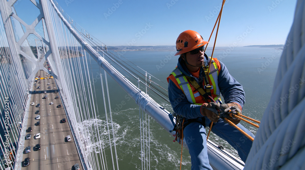 Fototapeta premium Construction Worker Ensures Safety in Bay Area Suspension Bridge Maintenance - Skilled Labor at Heights in Urban Engineering