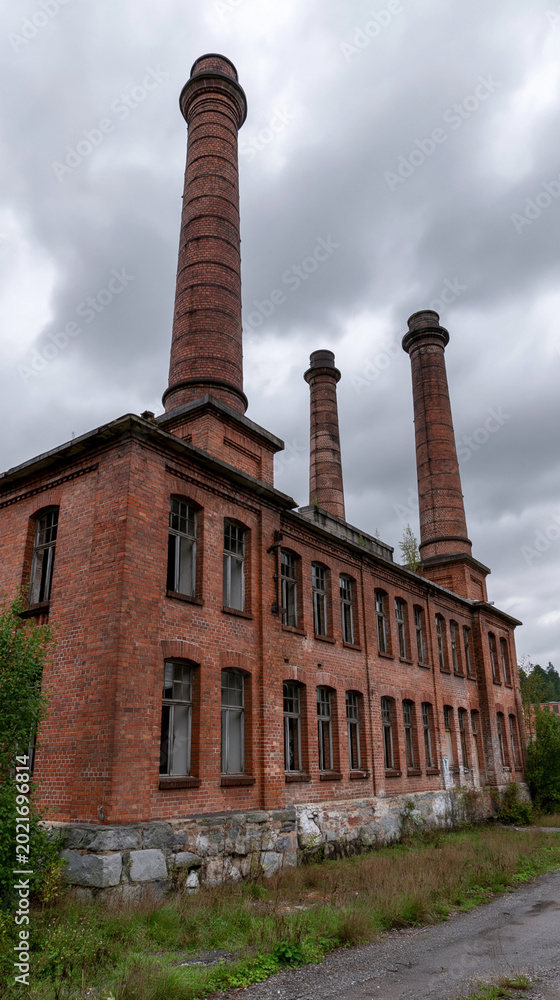 Fototapeta premium Historical Brick Factory: A striking image of a historic brick factory, its tall chimneys reaching towards a cloudy sky. The weathered facade whispers tales of a bygone industrial era.
