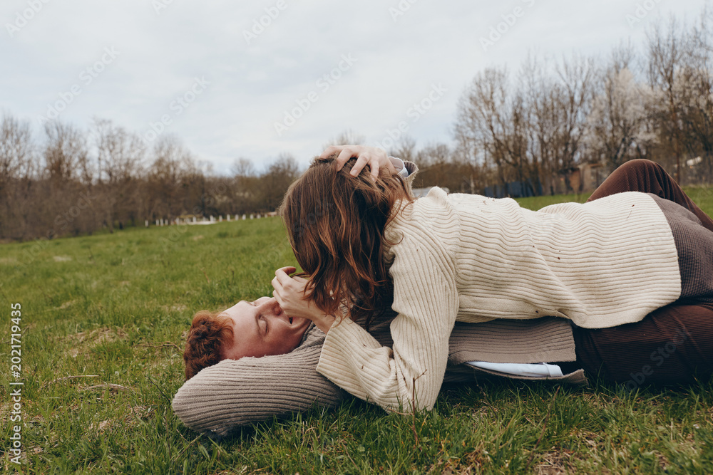 Fototapeta premium couple affection outdoors friendship laughter on the grass, a woman and a man share a playful moment in cozy sweaters, under a clear sky, showcasing casual lifestyle, outdoor leisure, and genuine