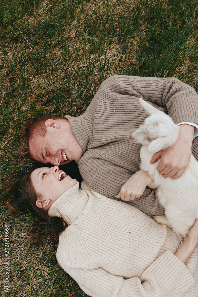 Fototapeta premium Smiling couple lying on grass with a curious goat kid, embracing and laughing in warm natural light. Cozy sweaters, outdoor vibe, intimate lifestyle moment with playful animal companionship.