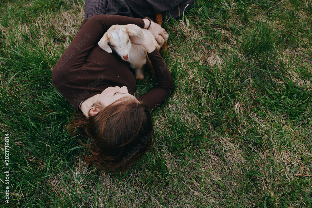 Fototapeta premium Relaxed woman lying on grass, cuddling a goat kid in a sunny field, capturing a calm outdoor lifestyle moment of affection, companionship, and simple nature joy.